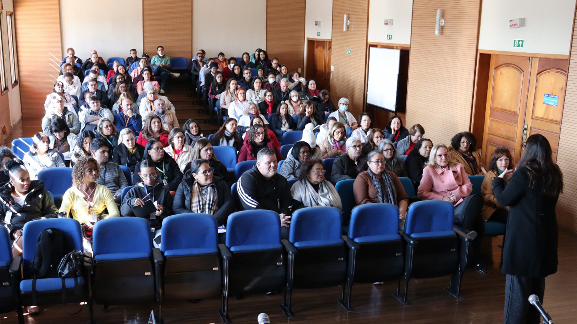Foto de uma sala cheio de pessoas sentadas, assistindo uma palestra.