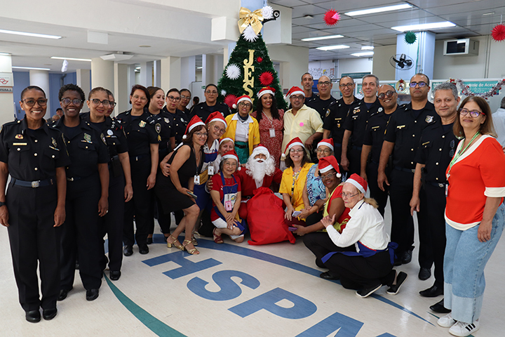 Foto colorida, em ambiente interno amplo, possivelmente um hospital ou instituição pública. Um grande grupo de pessoas adultas posa para a foto, de frente para a câmera, sorrindo. Ao centro há uma árvore de Natal decorada, com enfeites coloridos e a palavra “JESUS” escrita verticalmente. Em volta da árvore, há homens e mulheres de diferentes idades e tons de pele. Muitos usam uniformes pretos, organizados em fileiras laterais. Na parte central e à frente, algumas pessoas vestem roupas coloridas e gorros vermelhos de Natal. Uma pessoa caracterizada de Papai Noel está sentada no centro, usando roupa vermelha e barba branca. O chão tem uma grande marcação com letras azuis. O ambiente é bem iluminado e decorado de forma simples para o Natal. A cena transmite confraternização, alegria e clima de celebração natalina.