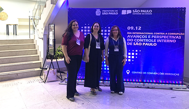Foto em ambiente interno de um centro de convenções. Em primeiro plano, três mulheres adultas estão de pé lado a lado, sorrindo e olhando para a câmera. Todas usam crachás de identificação pendurados no pescoço. À esquerda, há uma escada ampla que sobe para o piso superior. Ao fundo, à direita, aparece um grande painel iluminado em tons de azul e roxo. No painel, lê-se: “Dia Internacional Contra a Corrupção — Avanços e Perspectivas do Controle Interno de São Paulo”, com logotipos da Prefeitura de São Paulo e do Governo do Estado. O local é bem iluminado e transmite clima de evento institucional.