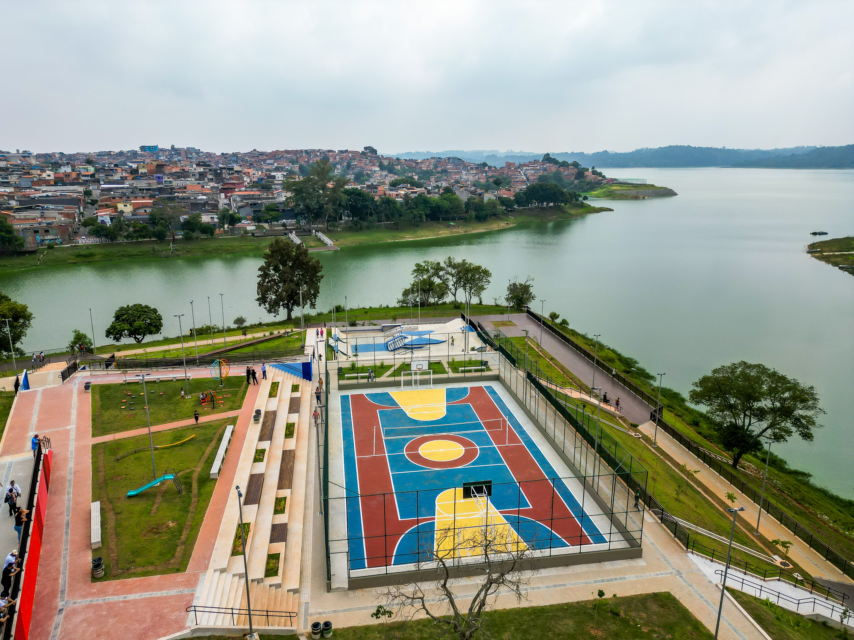 Vista aérea do Parque Pabreu Prainha com quadra colorida, pista de caminhada e área verde à beira da represa Billings. Ao fundo, um bairro com muitas casas.