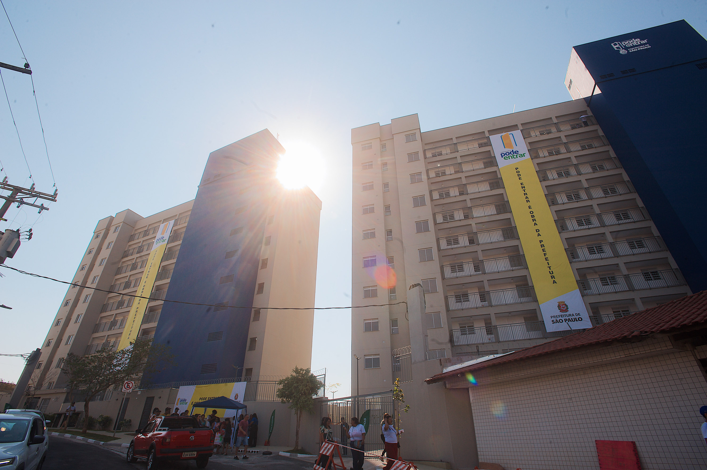 Dois prédios nas cores bege, cinza e azul, com pessoas em frente. Se estendendo nos prédios uma faixa comprida com a logo do programa Pode Entrar, uma porta amarela entreaberta sob um arco azul com a escrita pode entrar, ao longo da faixa está escrito 