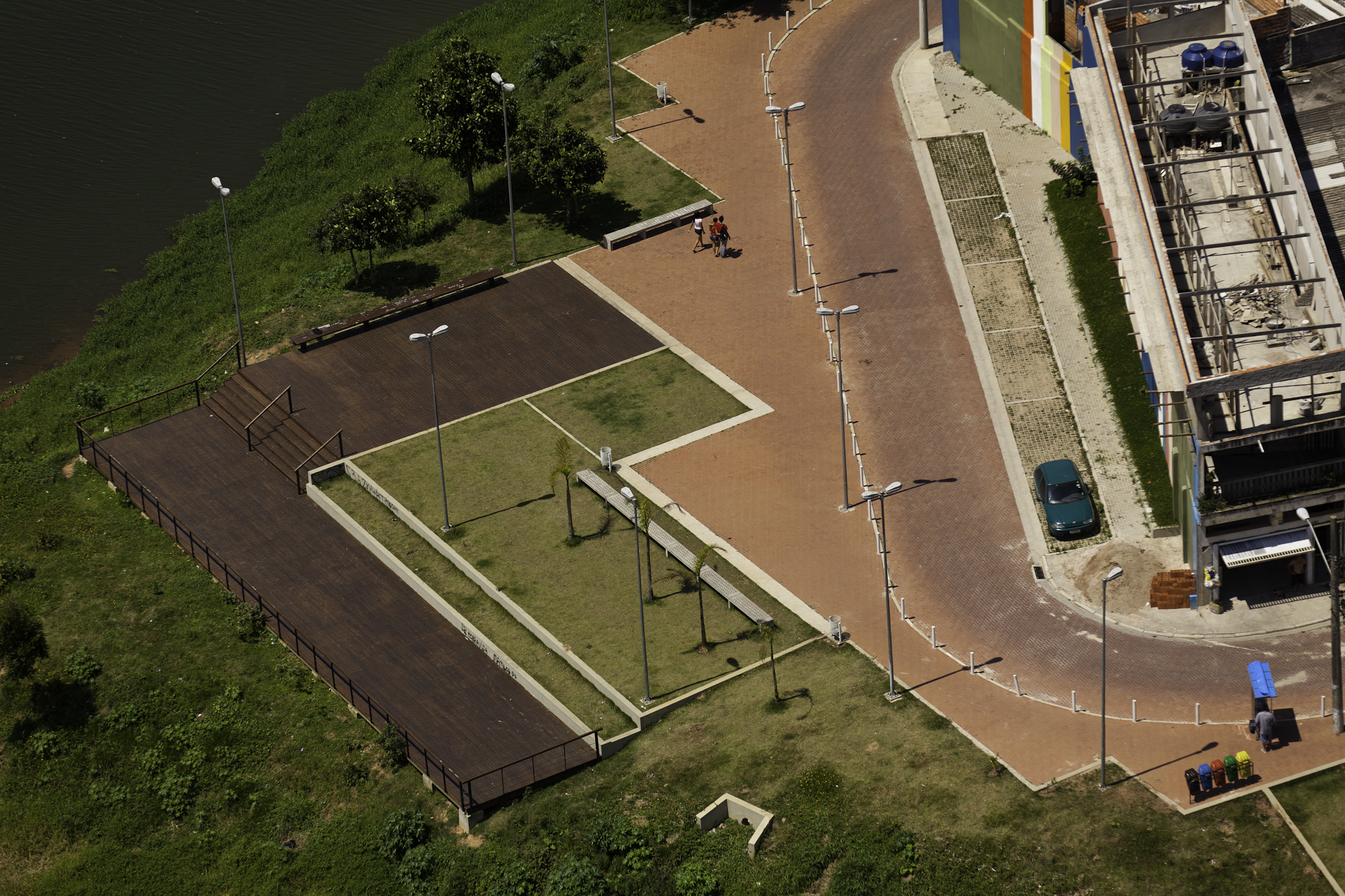Um espaço público visto de cima na região do Cantinho do Ceu, com calçadas largas, uma área gramada com árvores pequenas, bancos, postes de luz e um deck de madeira próximo a um rio.