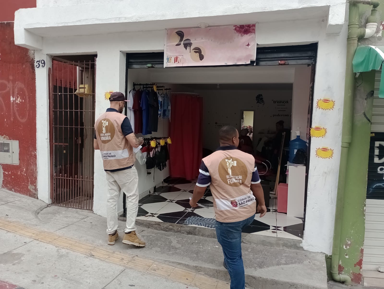 Dois homens de coletes bege da Regularização Fundiária – Cidade de São Paulo caminham pela calçada em direção à entrada de um pequeno comércio de roupas e salão de beleza. O espaço tem piso preto e branco em formato de losangos e uma cortina vermelha ao fundo. Na parede externa branca há adesivos amarelos em forma de sol e, ao lado esquerdo, o número 39 em azul. Um dos homens usa boné, máscara e botas claras, o outro veste calça jeans e tênis escuros.