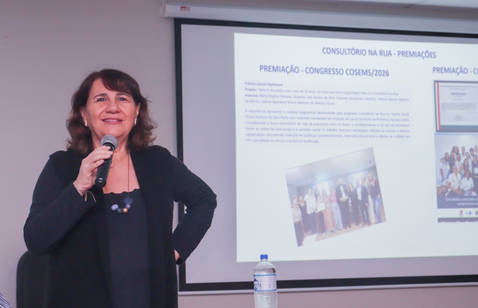 A imagem mostra uma mulher em pé, segurando um microfone e falando em uma apresentação ou palestra. Ela sorri para a câmera enquanto mantém uma das mãos apoiada na cintura. Está vestida com roupa preta e usa um colar discreto.  Ao fundo, há uma tela de projeção exibindo um slide com o título “Consultório na Rua – Premiações”. O slide parece apresentar informações sobre uma premiação recebida em um congresso, incluindo texto e pequenas fotografias de grupos de pessoas.  Na parte inferior da imagem, sobre uma mesa, há uma garrafa de água. O ambiente é interno, com iluminação suave, típico de auditórios ou salas de reunião usadas para eventos acadêmicos ou institucionais.