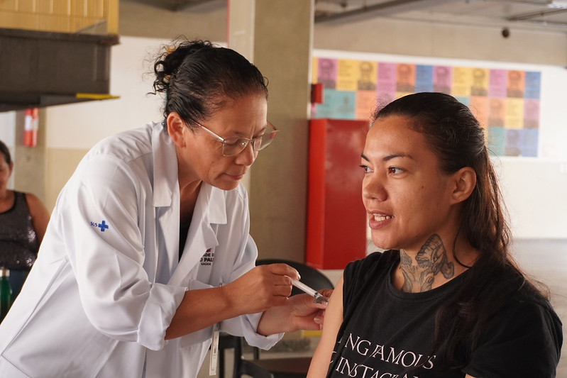 Profissional de saúde, uma mulher de jaleco branco, aplica uma vacina no braço esquerdo de outra mulher, que está sentada e usa camiseta preta. A pessoa vacinada olha para frente e mantém o braço relaxado, enquanto a profissional segura a seringa com atenção. O atendimento acontece em um espaço interno amplo, com pilares e outras pessoas ao fundo, indicando uma ação de vacinação em ambiente coletivo. 
