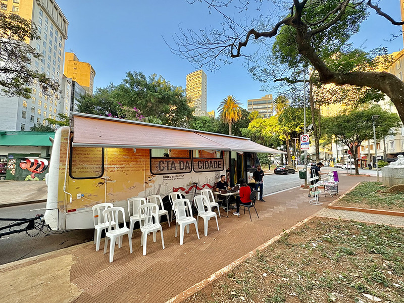 A imagem mostra uma unidade móvel de atendimento instalada em uma praça ou área pública arborizada de uma região central da cidade.  Em destaque está um trailer adaptado, identificado como “CTA da Cidade”, sigla normalmente associada aos Centros de Testagem e Aconselhamento em saúde. O veículo possui uma cobertura retrátil aberta sobre a calçada, criando uma área de atendimento ao público. Em frente ao trailer há várias cadeiras plásticas brancas organizadas para espera, enquanto algumas pessoas conversam ou realizam atendimentos em mesas instaladas ao lado da unidade.  O cenário urbano é composto por prédios altos ao fundo, árvores de grande porte, palmeiras e uma praça com áreas de circulação de pedestres. A luz do sol ilumina a cena, sugerindo que a atividade ocorre durante o dia