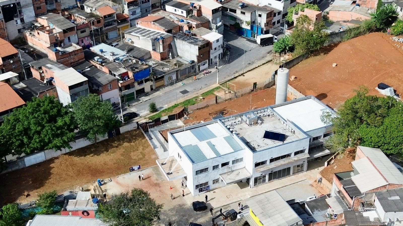 A imagem apresenta uma vista aérea, em ângulo zenital, de uma área urbana densamente povoada. O foco central da imagem é um prédio de dois andares com arquitetura moderna e acabamento em tons claros, que está em fase final de construção ou acabamento. O edifício possui um telhado plano com equipamentos de climatização e painéis solares instalados. Ao redor do prédio, há uma área de terreno exposto, indicando o canteiro de obras, e é possível identificar algumas pessoas circulando na parte inferior. O entorno é caracterizado por uma malha de casas residenciais geminadas, estreitas e com telhados de cerâmica, típicas de periferias urbanas. Uma rua asfaltada contorna o terreno, onde se percebe um fluxo leve de veículos e pedestres.