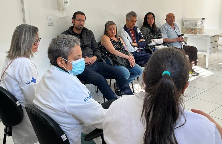 A imagem mostra uma atividade coletiva realizada em uma unidade de saúde, reunindo profissionais e usuários em uma roda de conversa.  Em primeiro plano, três profissionais da saúde, vestindo jalecos brancos com a identificação do SUS, participam da discussão. Um deles utiliza máscara cirúrgica. À frente deles, sentados em cadeiras dispostas em círculo, estão homens e mulheres de diferentes faixas etárias, incluindo pessoas idosas, acompanhando atentamente o encontro.  O ambiente é claro, organizado e bem iluminado, com paredes brancas e mobiliário simples. A disposição das cadeiras em círculo favorece a interação e o diálogo entre participantes e equipe de saúde.  A cena remete a ações de promoção da saúde, educação em saúde ou grupos terapêuticos desenvolvidos na Atenção Básica, em que usuários compartilham experiências, recebem orientações e fortalecem vínculos com os profissionais e com a comunidade. A fotografia transmite uma ideia de acolhimento, escuta qualificada e cuidado coletivo, características importantes do trabalho realizado nas Unidades Básicas de Saúde (UBSs).