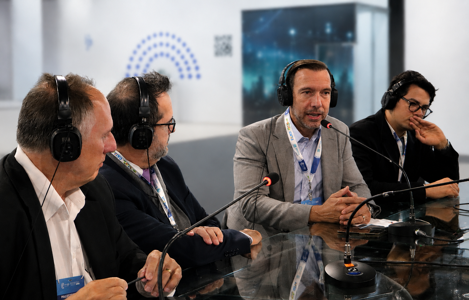 A imagem mostra uma mesa-redonda ou entrevista realizada em um ambiente de feira ou congresso corporativo. Em primeiro plano, quatro homens estão sentados ao redor de uma mesa de vidro, utilizando fones de ouvido e microfones, em uma conversa ou debate transmitido ao vivo. No centro da cena, um dos participantes, vestido com blazer cinza claro e camisa social, fala ao microfone enquanto mantém as mãos apoiadas sobre a mesa. Os demais participantes, trajando ternos escuros, voltam sua atenção para ele, demonstrando interesse e escuta ativa. A mesa possui acabamento espelhado, refletindo os participantes, os microfones e parte dos crachás utilizados durante o evento. Os equipamentos de áudio reforçam o caráter profissional da atividade, sugerindo uma gravação de podcast, entrevista ou painel de discussão. A composição transmite uma atmosfera de diálogo especializado, networking e troca de conhecimentos em um contexto de inovação, tecnologia ou negócios.