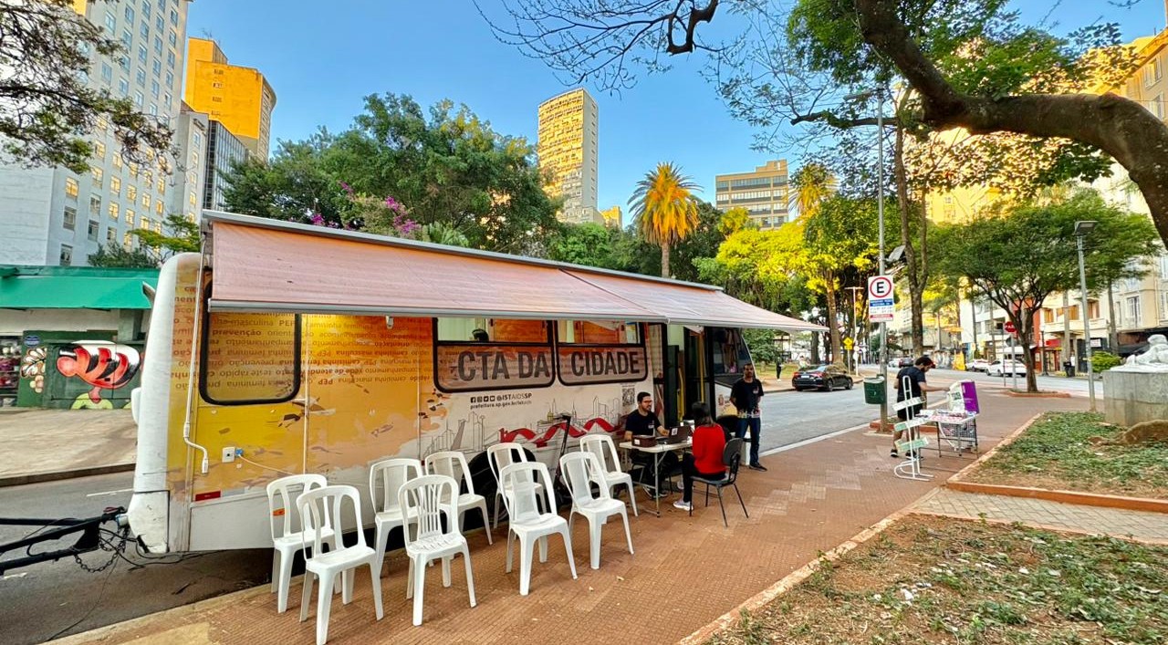 A imagem mostra uma unidade móvel de atendimento instalada em uma praça ou área pública arborizada de uma região central da cidade.  Em destaque está um trailer adaptado, identificado como “CTA da Cidade”, sigla normalmente associada aos Centros de Testagem e Aconselhamento em saúde. O veículo possui uma cobertura retrátil aberta sobre a calçada, criando uma área de atendimento ao público. Em frente ao trailer há várias cadeiras plásticas brancas organizadas para espera, enquanto algumas pessoas conversam ou realizam atendimentos em mesas instaladas ao lado da unidade.  O cenário urbano é composto por prédios altos ao fundo, árvores de grande porte, palmeiras e uma praça com áreas de circulação de pedestres. A luz do sol ilumina a cena, sugerindo que a atividade ocorre durante o dia.