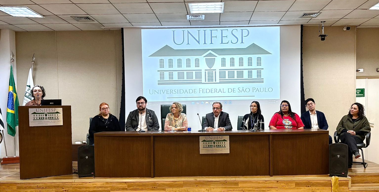 A imagem mostra uma mesa de debate ou evento acadêmico realizado em um auditório da Universidade Federal de São Paulo.  Ao fundo, há uma grande tela de projeção exibindo o logotipo e o nome da universidade. Na frente da sala, sete pessoas estão sentadas atrás de uma longa mesa de madeira, posicionadas lado a lado, enquanto outra mulher fala em um púlpito à esquerda da imagem.  A palestrante está em pé atrás do púlpito, utilizando um microfone e um computador. Ao lado dela aparecem as bandeiras do Brasil e da instituição. Os participantes da mesa usam roupas variadas, desde trajes sociais até roupas mais casuais, sugerindo um encontro institucional ou discussão temática.  O auditório possui iluminação clara, teto com painéis e um ambiente formal. Na parte frontal da mesa há placas com o nome da universidade, reforçando o caráter oficial e acadêmico do evento.