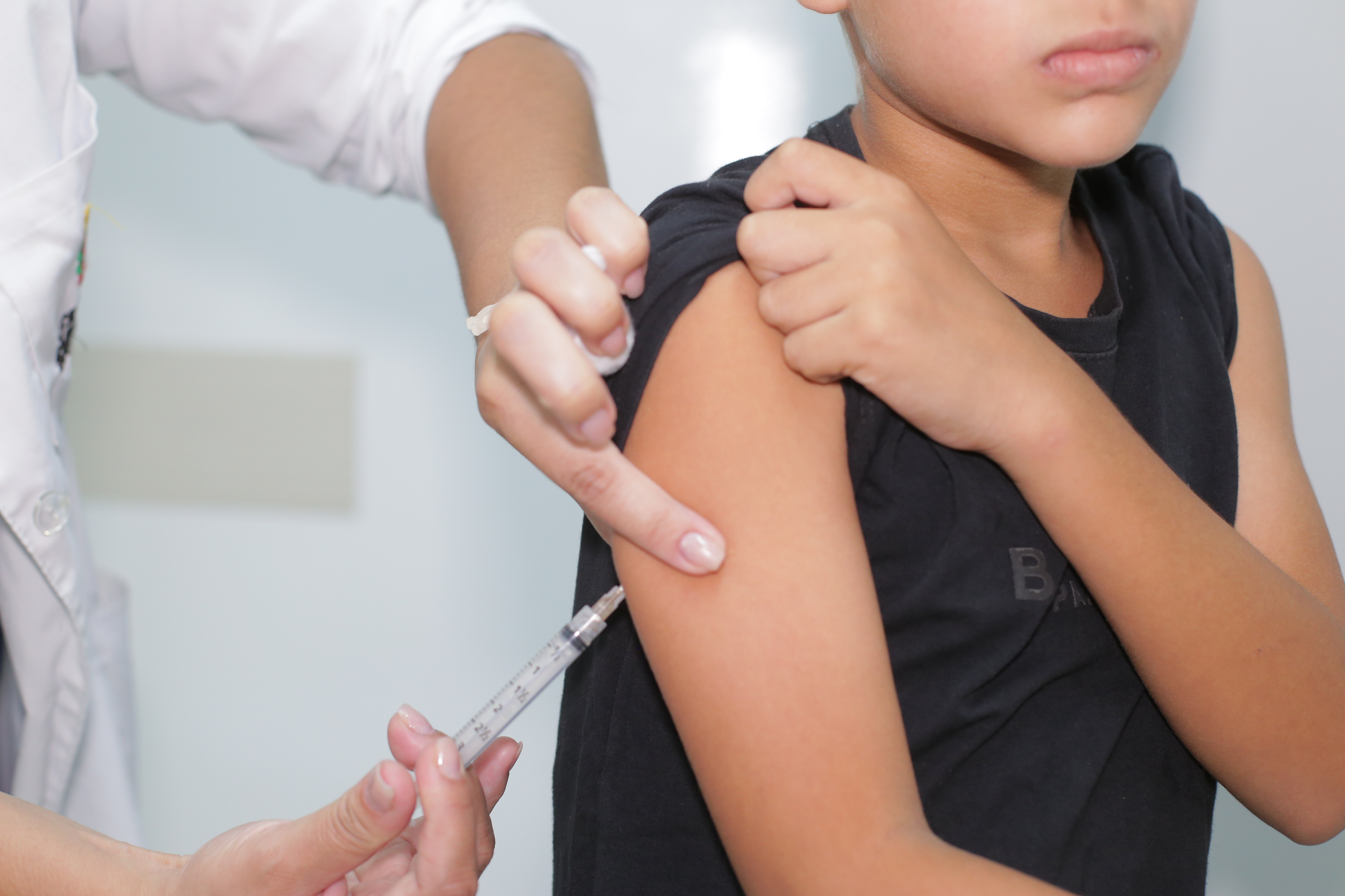 A imagem mostra o momento em que uma criança recebe uma vacina no braço esquerdo, durante um atendimento em uma unidade de saúde.  Em primeiro plano, um profissional de saúde, vestido com jaleco branco, prepara a aplicação da vacina com uma seringa, enquanto utiliza uma das mãos para estabilizar o braço da criança e indicar o local da injeção. A criança segura a manga da camiseta preta para deixar o ombro descoberto e mantém uma expressão séria durante o procedimento.  A fotografia enquadra apenas parte do rosto da criança e das mãos do profissional, direcionando a atenção para o ato da vacinação. O fundo desfocado reforça o ambiente clínico e destaca o procedimento.
