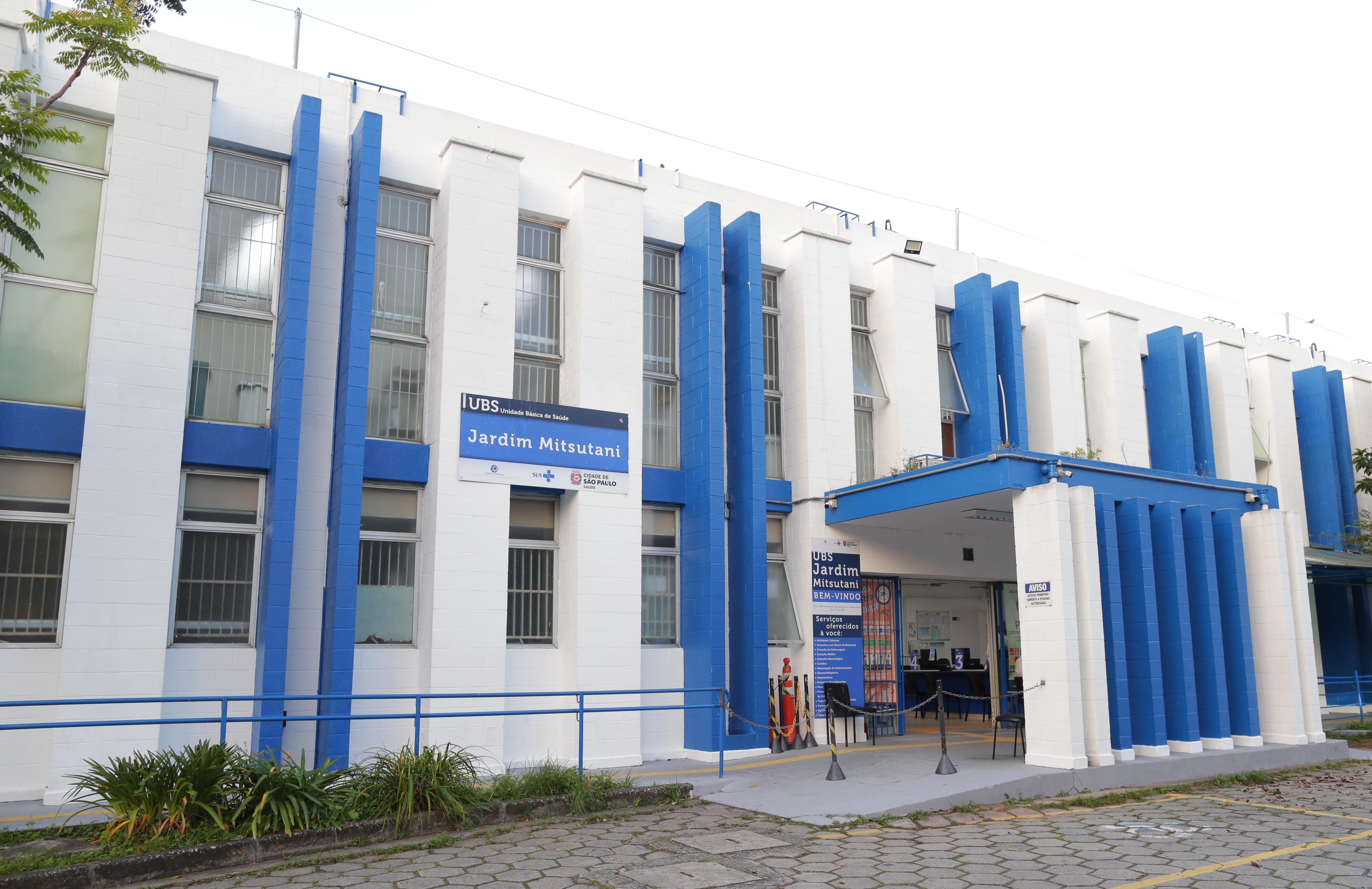 A imagem mostra a fachada da UBS Jardim Mitsutani, uma Unidade Básica de Saúde localizada na cidade de São Paulo. O prédio é de dois andares, com pintura predominantemente branca e detalhes em azul, transmitindo aparência organizada e bem conservada.  Na entrada, há uma marquise azul com o nome da unidade destacado em uma placa que traz também o logotipo da Prefeitura de São Paulo e do SUS (Sistema Único de Saúde). O local possui acesso adaptado com rampa e corrimão, além de uma área externa pavimentada e pequenas plantas ao redor. O interior da unidade é parcialmente visível, com balcões de atendimento e cartazes informativos, indicando um ambiente voltado ao acolhimento e orientação dos usuários do serviço público de saúde.