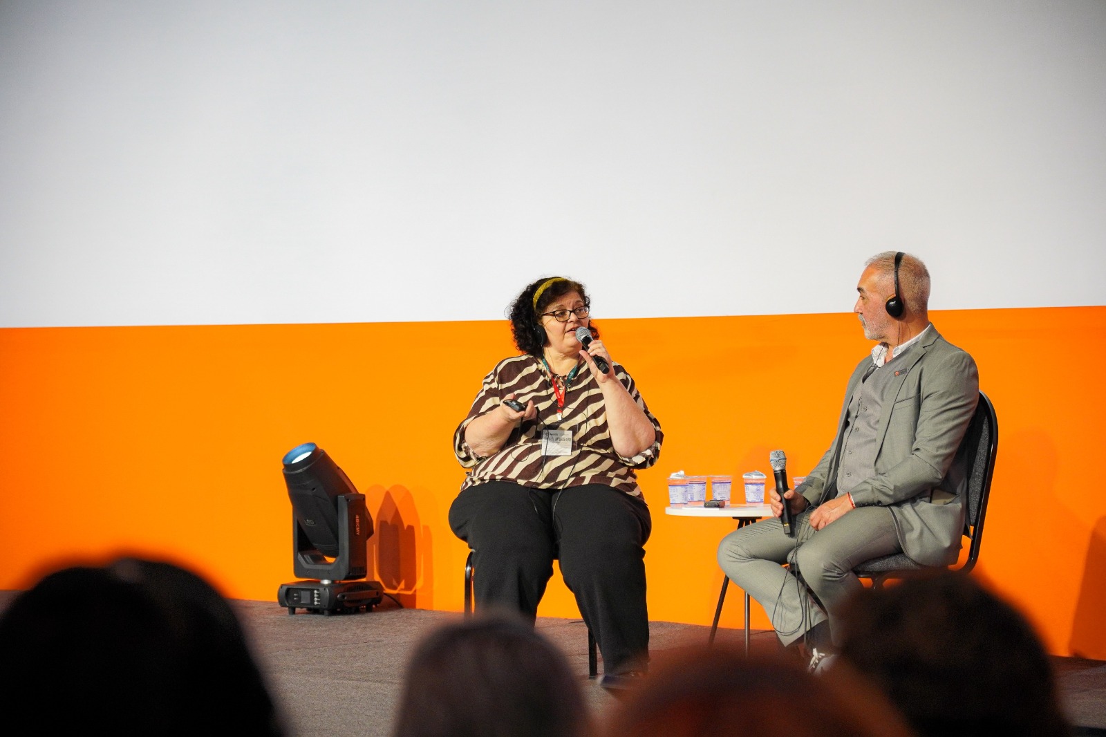 A imagem mostra duas pessoas sentadas em cadeiras no palco de um auditório, durante a ExpoLongevidade. À esquerda, uma mulher de pele clara, cabelos curtos e cacheados de cor preta, usa uma faixa amarela no cabelo e óculos de armação escura. Ela veste uma blusa com estampa geométrica em tons de marrom e bege e calça preta. Segura um microfone em uma das mãos e parece estar falando.  À direita, um homem de pele clara e cabelos grisalhos, curtos, também usa fones de ouvido e segura um microfone. Ele veste um terno cinza-claro com camisa branca. Entre eles, há uma pequena mesa redonda com copos e garrafas de água.  O fundo é dividido em duas cores: a parte superior branca e a inferior laranja. No chão, há um refletor preto apontado para o palco. O público aparece parcialmente desfocado na parte inferior da imagem.