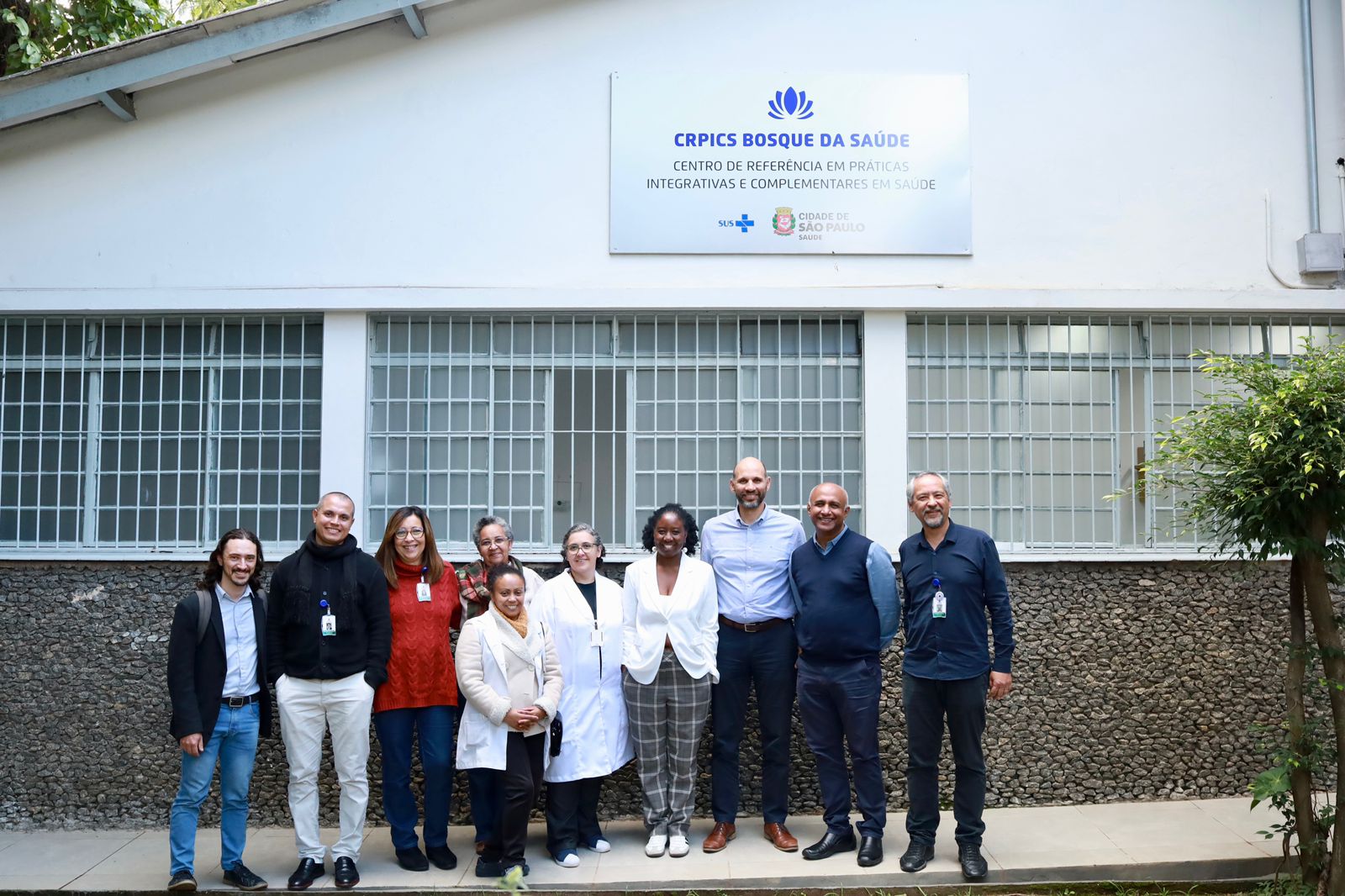 Grupo de Profissionais de Saúde estão em frente a Sede do Centro de Referência em Práticas Integrativas e Complementares em Saúde. Uma placa acima destaca as iniciais do equipamento CRPICS Bosque da Saúde.   