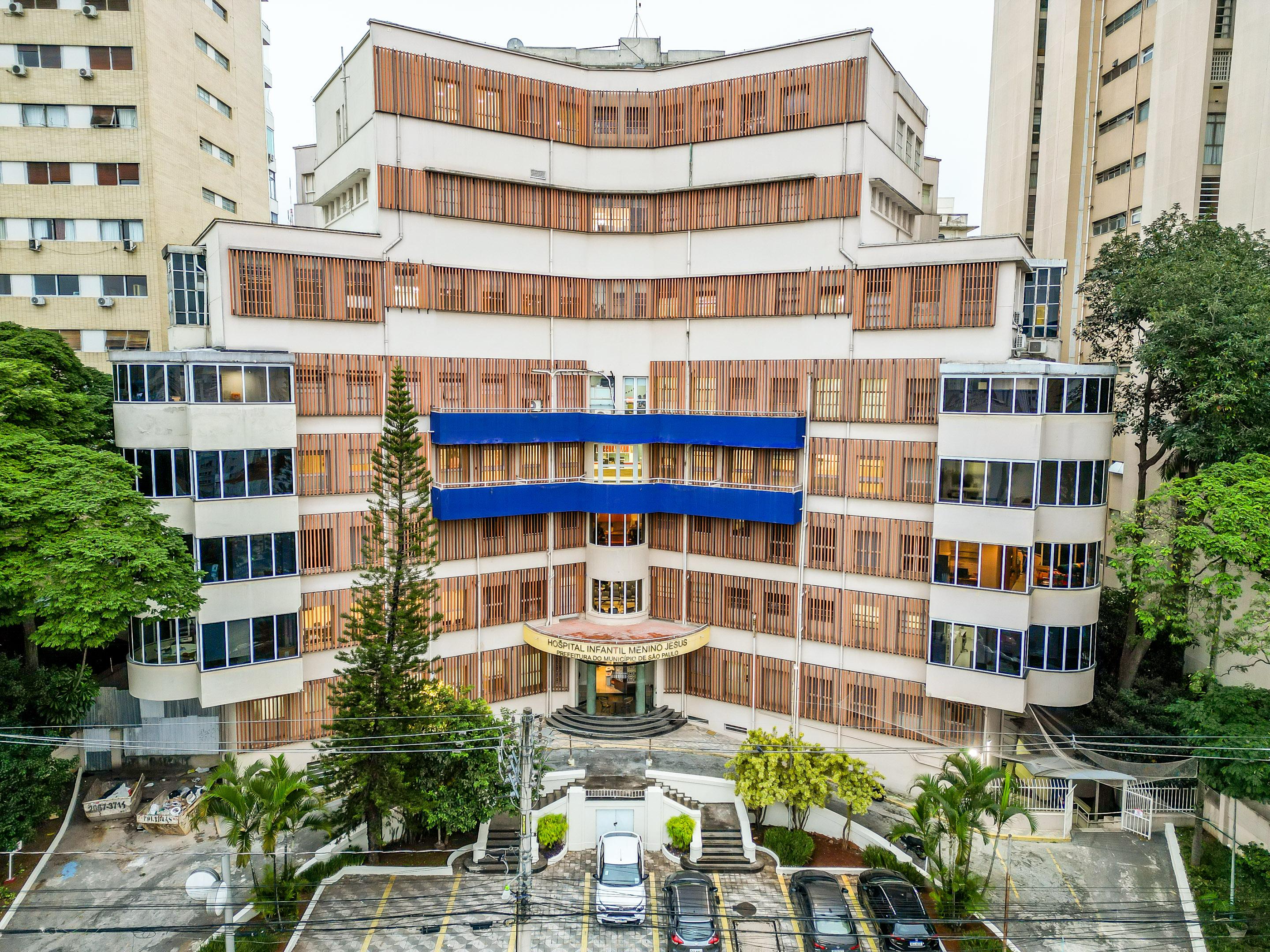 A imagem mostra a fachada do Hospital Infantil Menino Jesus, localizado em São Paulo. O prédio é de arquitetura moderna e simétrica, com várias janelas e estruturas verticais de madeira que funcionam como brises. Na parte central do edifício, destacam-se duas faixas horizontais azuis que percorrem os andares médios da construção. O letreiro acima da entrada principal identifica claramente o local como “Hospital Infantil Menino Jesus”. Há uma escadaria semicircular que leva até a porta principal, ladeada por canteiros com vegetação. Em frente ao prédio, vê-se um pequeno estacionamento com carros estacionados e fios de eletricidade atravessando a imagem. O hospital está cercado por outros edifícios altos e árvores, compondo um ambiente urbano e arborizado.