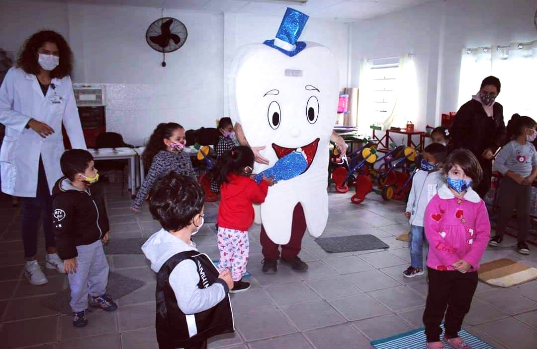 A imagem mostra um grupo de crianças participando de uma atividade educativa em um ambiente escolar. No centro, há uma pessoa vestida com uma fantasia grande e branca em forma de dente, usando um chapéu azul brilhante e um laço da mesma cor, simbolizando o cuidado com a saúde bucal. As crianças, que usam máscaras coloridas, parecem atentas e animadas com a presença do personagem. À esquerda, uma mulher de jaleco branco, possivelmente uma profissional de saúde, observa a interação. Ao fundo, há brinquedos e triciclos, indicando que a atividade acontece em uma sala infantil. A cena transmite alegria e aprendizado sobre higiene bucal.