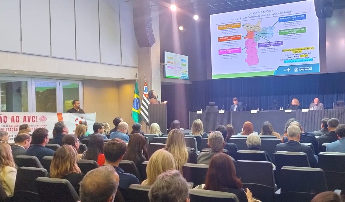 A foto mostra um auditório cheio, com dezenas de pessoas sentadas assistindo a uma apresentação. À frente, há uma mesa com quatro pessoas — três sentadas e uma mulher em pé, falando ao microfone e apontando para um telão. No telão é exibido um mapa da cidade de São Paulo, com a divisão por Coordenadorias Regionais de Saúde e informações populacionais. Ao lado do palco estão as bandeiras do Brasil e do estado de São Paulo. À esquerda, um banner com os dizeres “Não ao AVC!” é segurado por um homem próximo à parede. O ambiente tem iluminação clara e aspecto institucional, indicando um evento oficial, possivelmente ligado à Secretaria Municipal de Saúde