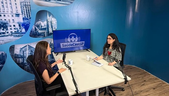 A imagem mostra duas mulheres sentadas frente a frente em um estúdio de gravação, participando de uma conversa ou entrevista em formato de podcast. Elas estão posicionadas em uma mesa branca, cada uma com um microfone profissional à sua frente, indicando um ambiente preparado para captação de áudio.  Ao fundo, há um painel azul com o logotipo “SaúdeCastSP” exibido em uma tela, além de imagens urbanas que remetem à cidade de São Paulo. O espaço é bem iluminado, organizado e com aparência institucional, sugerindo um programa voltado à área da saúde ou a temas relacionados a políticas públicas e serviços.  Uma das participantes parece conduzir a conversa, gesticulando enquanto fala, enquanto a outra escuta atentamente, com anotações sobre a mesa. O cenário transmite profissionalismo e um clima de diálogo informativo.