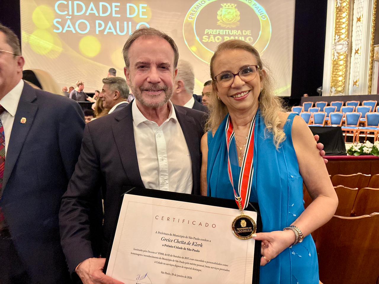 A imagem mostra duas pessoas posando para uma foto em um evento solene, aparentemente em uma cerimônia oficial na cidade de São Paulo.  À esquerda, há um homem vestindo terno escuro e camisa branca, sorrindo para a câmera. À direita, há uma mulher com cabelo claro preso, usando óculos e um vestido azul, também sorridente. Ela usa no pescoço uma medalha com fita tricolor, indicando que recebeu uma homenagem ou premiação.  Ambos seguram um certificado emoldurado, no qual é possível ler “Certificado” e “Prêmio Cidade de São Paulo”, concedido pela Prefeitura. O documento indica que se trata de uma homenagem oficial.  Ao fundo, vê-se um palco com cadeiras organizadas, além de um telão com os dizeres “Cidade de São Paulo” e o brasão da prefeitura, reforçando o contexto institucional do evento.  A cena transmite um clima de reconhecimento, celebração e prestígio, típico de uma cerimônia de premiação pública.
