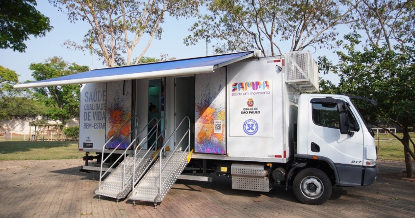 A imagem mostra uma unidade móvel de saúde da Prefeitura de São Paulo estacionada em um ambiente externo arborizado. O veículo é um caminhão adaptado com um toldo azul e duas entradas acessíveis por escadas metálicas. Na lateral do caminhão, estão estampadas as palavras 