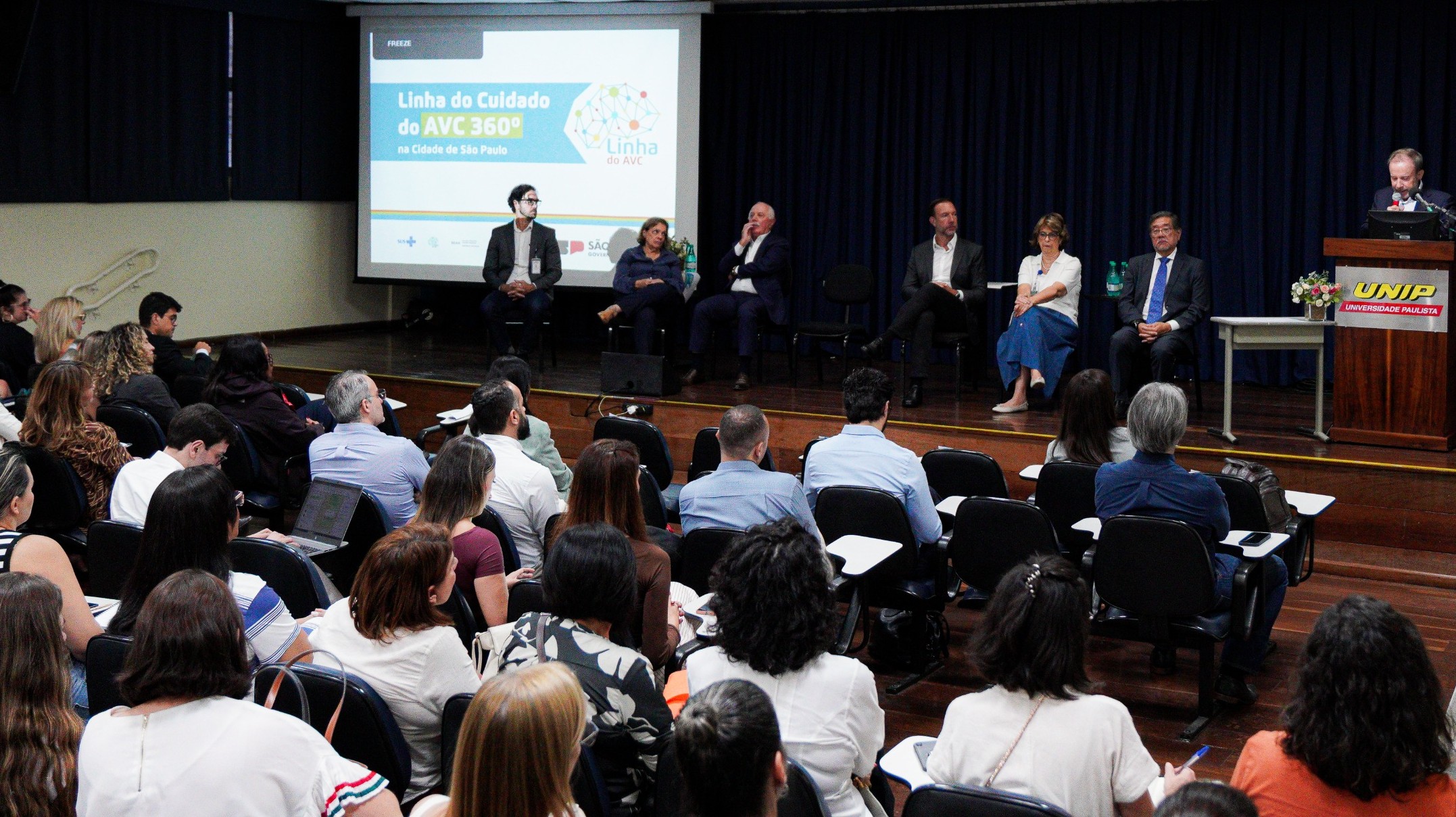 A imagem mostra um auditório lotado durante um evento ou seminário na área da saúde. No palco, há uma mesa com vários especialistas sentados, enquanto um homem fala ao púlpito, identificado com o logotipo da UNIP (Universidade Paulista).  Ao fundo, um telão exibe a apresentação “Linha do Cuidado do AVC 360º na Cidade de São Paulo”, indicando que o encontro aborda a organização e o atendimento integral aos casos de Acidente Vascular Cerebral.  Na plateia, composta majoritariamente por adultos, profissionais da área da saúde acompanham atentamente a discussão, alguns fazendo anotações ou utilizando notebooks. As cadeiras estão organizadas em fileiras, voltadas para o palco.  O ambiente é formal e institucional, característico de um evento técnico ou científico, voltado à capacitação, troca de experiências e fortalecimento das políticas públicas de saúde.