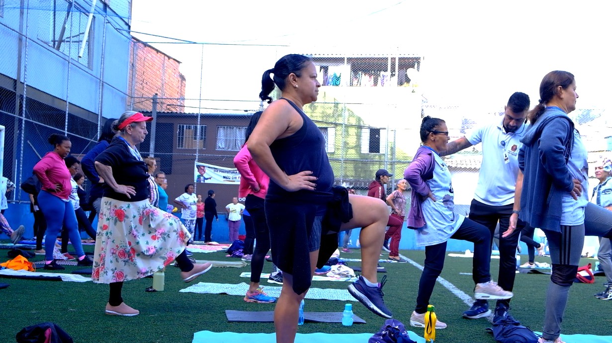 A imagem mostra um grupo de mulheres participando de uma atividade física ao ar livre, em uma quadra esportiva cercada por prédios residenciais. Elas estão organizadas em fileiras, sobre tapetes ou toalhas no chão, realizando um exercício de equilíbrio, com uma perna apoiada no solo e a outra elevada. A maioria usa roupas confortáveis para prática esportiva, como leggings, camisetas e tênis. Algumas participantes vestem uniformes claros, possivelmente de um programa comunitário ou de saúde. À direita, um profissional, provavelmente um educador físico ou agente de saúde, orienta os movimentos e auxilia uma das participantes. No fundo, outras pessoas observam ou também participam da atividade. O ambiente sugere uma ação comunitária voltada à promoção da saúde, bem-estar e convivência, em um espaço público do bairro.