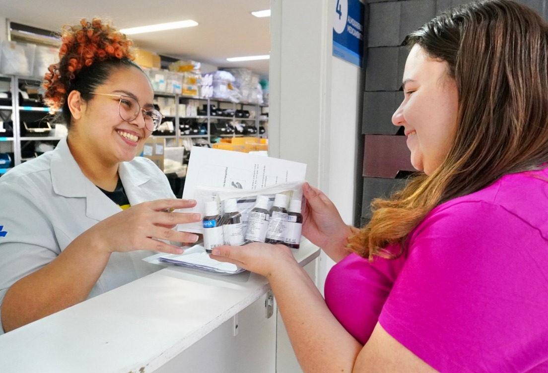 ​​​​​​​A imagem captura um momento de interação positiva em um ambiente de saúde ou farmácia. A foto mostra duas mulheres em um balcão de atendimento. A iluminação é clara e direta, típica de ambientes profissionais ou clínicos. Ao fundo, é possível ver estantes organizadas com diversas caixas e suprimentos, sugerindo que o local seja uma farmácia hospitalar, um almoxarifado de medicamentos ou uma unidade de saúde