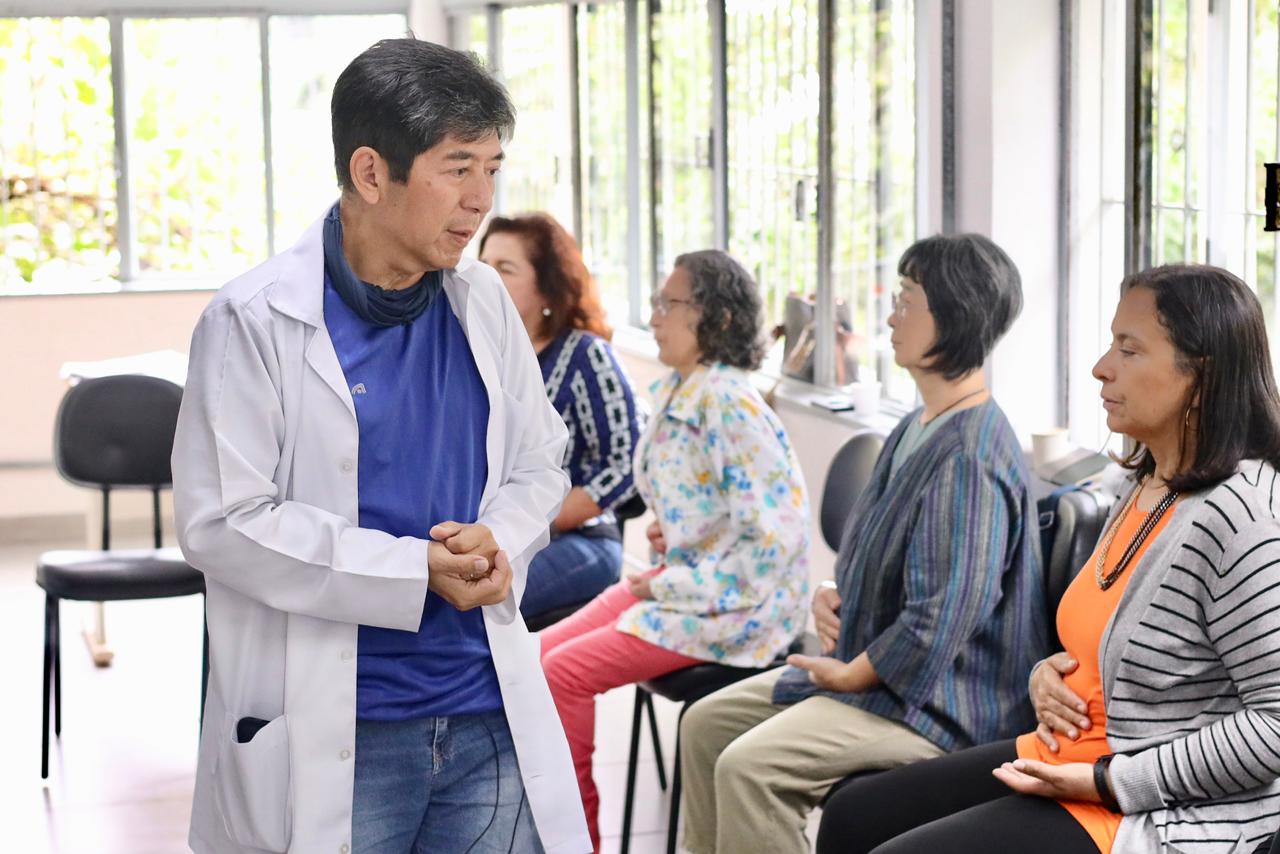 A foto mostra um ambiente interno iluminado por janelas amplas ao fundo. Em primeiro plano, à esquerda, um homem de traços asiáticos, cabelos curtos e escuros, veste um jaleco branco sobre uma camiseta azul e calça jeans. Ele está em pé, com as mãos unidas à frente do corpo, em uma postura de quem explica algo com atenção.  À direita, quatro mulheres estão sentadas em cadeiras individuais, formando uma fila ou círculo de conversa. Elas parecem praticar um exercício de respiração consciente ou meditação, com as mãos repousadas sobre o abdômen e olhos fechados ou semicerrados. O clima é de tranquilidade, foco e aprendizado coletivo.