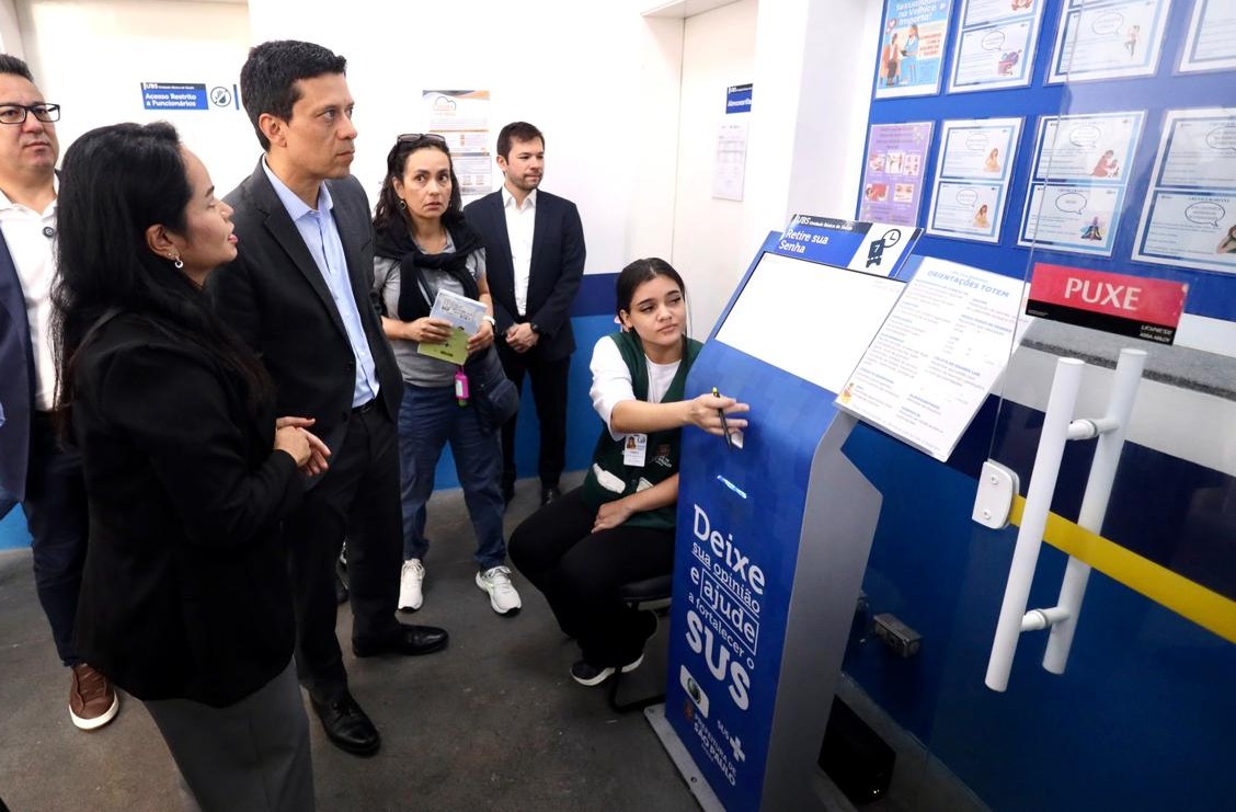 A imagem mostra um grupo de pessoas em uma unidade de saúde, aparentemente durante uma visita técnica ou institucional. Ao centro, um homem de terno observa atentamente uma funcionária sentada, que aponta para um equipamento de autoatendimento — possivelmente um totem de retirada de senha ou avaliação de serviços do SUS.  Ao redor, outras pessoas também acompanham a explicação, incluindo homens e mulheres em trajes formais e uma mulher com crachá, sugerindo que são gestores, técnicos ou visitantes. A funcionária veste uniforme e parece estar demonstrando o funcionamento do equipamento.  Na parede à direita, há um painel com o título “Cardápio de Grupos”, com materiais informativos organizados, provavelmente relacionados a atividades educativas ou serviços oferecidos pela unidade. O ambiente é limpo, bem iluminado e organizado, típico de um equipamento público de saúde.