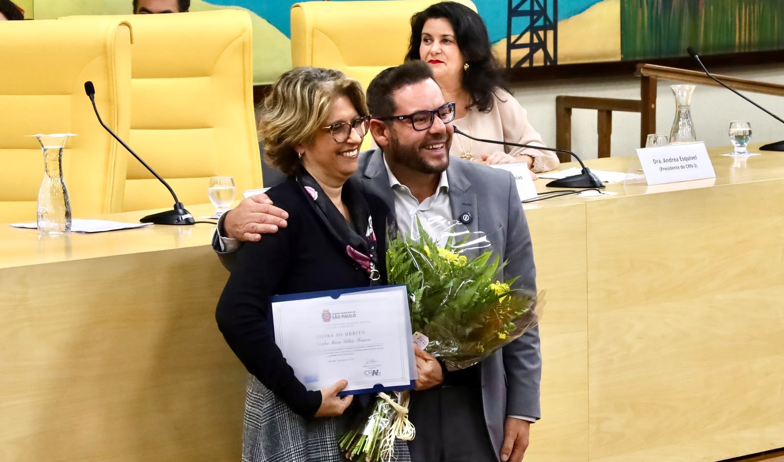 A imagem mostra um momento de homenagem em um auditório. Em primeiro plano, uma mulher e um homem aparecem sorrindo e posando juntos para a fotografia. A mulher segura um certificado de homenagem ao mérito e um buquê de flores, enquanto o homem a abraça pelo ombro.  Ao fundo, há uma mesa de cerimônia com microfones, copos e jarras de água, além de cadeiras amarelas. Atrás deles, uma terceira mulher está sentada à mesa, e é possível ver uma identificação com o nome “Dra. Andrea Esquivel” e a referência ao CRM. O ambiente tem características de uma solenidade institucional, com elementos decorativos e a identificação da Prefeitura de São Paulo visível no certificado.