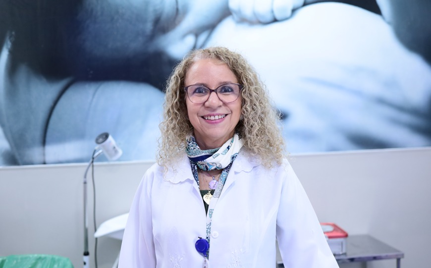 A imagem mostra uma médica em um consultório de ginecologia. No centro da foto está uma mulher em pé, olhando diretamente para a câmera e sorrindo. Ela tem cabelos loiros, longos e cacheados, usa e veste um jaleco branco. No pescoço, ela usa um lenço estampado e também um crachá preso ao jaleco. No consultório tem alguns equipamentos médicos sobre uma bancada, como uma luminária de exame e materiais sobre a mesa. Na parede atrás dela há uma grande imagem em tons de azul que mostra um bebê próximo ao corpo de uma mulher. A iluminação da imagem é clara e suave, destacando o rosto da profissional e criando um ambiente limpo e acolhedor.