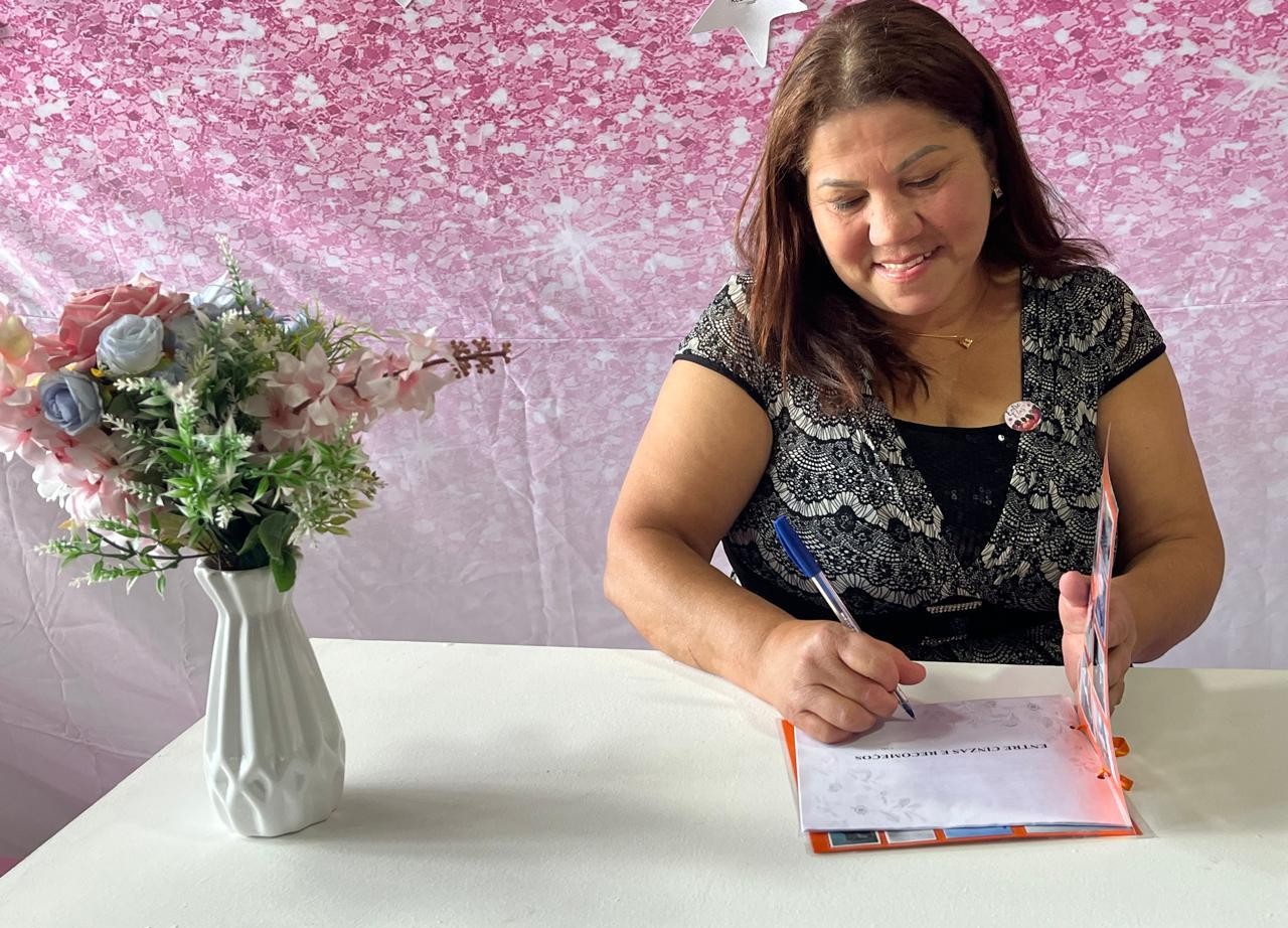 A imagem mostra uma mulher sentada à mesa durante uma sessão de autógrafos, assinando um exemplar de um livro. Ela sorri enquanto escreve na página de dedicatória com uma caneta azul. Está vestindo uma blusa preta com detalhes em renda branca sobre uma camiseta preta e usa um colar delicado e um broche na altura do peito. Sobre a mesa branca, à esquerda, há um vaso branco com um arranjo de flores artificiais em tons de rosa, lilás e branco, que ajuda a compor um ambiente acolhedor. Ao fundo, um painel decorativo em tons de rosa, com efeito de brilho e pequenas estrelas, reforça o caráter comemorativo da ocasião. O livro aberto revela o início do texto, com o título