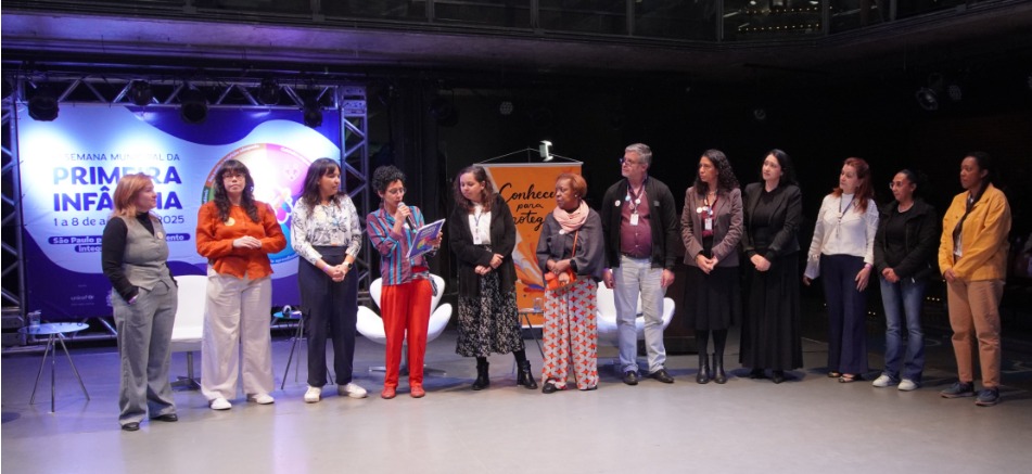 A imagem mostra um grupo de 12 pessoas enfileiradas em um palco, aparentemente participando de um evento oficial. No fundo, há um grande painel iluminado com a inscrição “Semana Municipal da Primeira Infância – 1 a 8 de agosto de 2025”, com o brasão da cidade de São Paulo e menção ao “Plano Municipal da Primeira Infância”. À direita do palco, há um banner laranja com a frase “Conhecer para proteger” e a imagem estilizada de uma criança. O grupo é diverso, formado por mulheres e um homem, com trajes formais ou casuais. Uma das mulheres, ao centro, está com um microfone e lendo um papel, indicando que é a pessoa que fala no momento. O clima é de solenidade ou abertura de um evento institucional relacionado à infância, com foco em políticas públicas, proteção e desenvolvimento infantil.