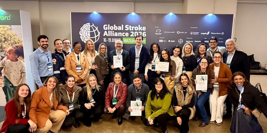 A foto mostra um grande grupo de pessoas reunidas em um evento, posando de forma descontraída e comemorativa diante de um painel institucional.  Ao fundo, há um banner com a inscrição “Global Stroke Alliance 2026”, além das datas do evento e diversos logotipos de organizações apoiadoras. O grupo é composto por homens e mulheres de diferentes idades, muitos usando crachás de identificação pendurados no pescoço, indicando participação em um congresso ou encontro profissional.  As pessoas estão distribuídas em duas fileiras: algumas em pé e outras agachadas na parte da frente. Vários participantes levantam os braços e acenam para a câmera, transmitindo entusiasmo, integração e celebração. Algumas pessoas seguram certificados ou placas de reconhecimento, sugerindo uma premiação, apresentação de projetos ou destaque em atividades do evento.  O ambiente é interno, com iluminação clara e profissional. As expressões são predominantemente sorridentes e alegres, reforçando o clima de confraternização e conquista coletiva. No canto inferior direito, é possível ver parte de um celular fotografando a mesma cena, indicando que o momento estava sendo registrado por diferentes participantes.