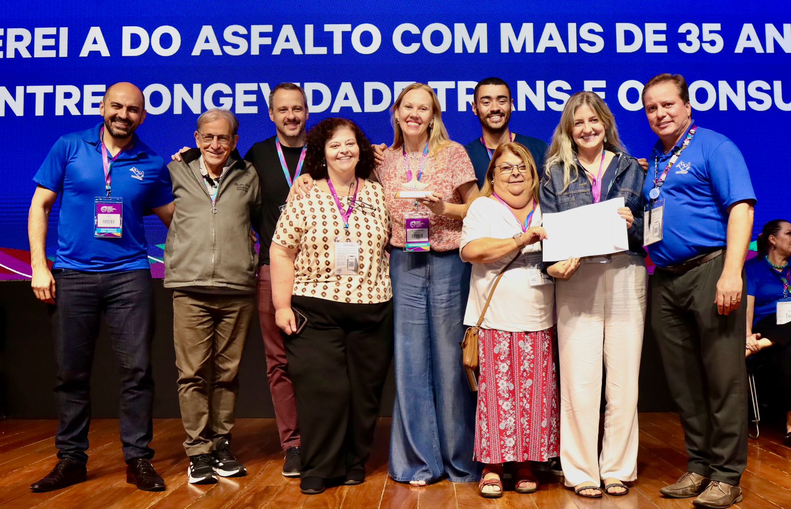 Grupo de dez pessoas posa lado a lado sobre um palco, sorrindo para a foto, durante um evento. Ao centro, uma mulher segura um troféu e, à direita, outra participante exibe um certificado. Todos usam crachás e estão diante de um painel azul com letras grandes ao fundo. A cena transmite um momento de reconhecimento e celebração coletiva.
