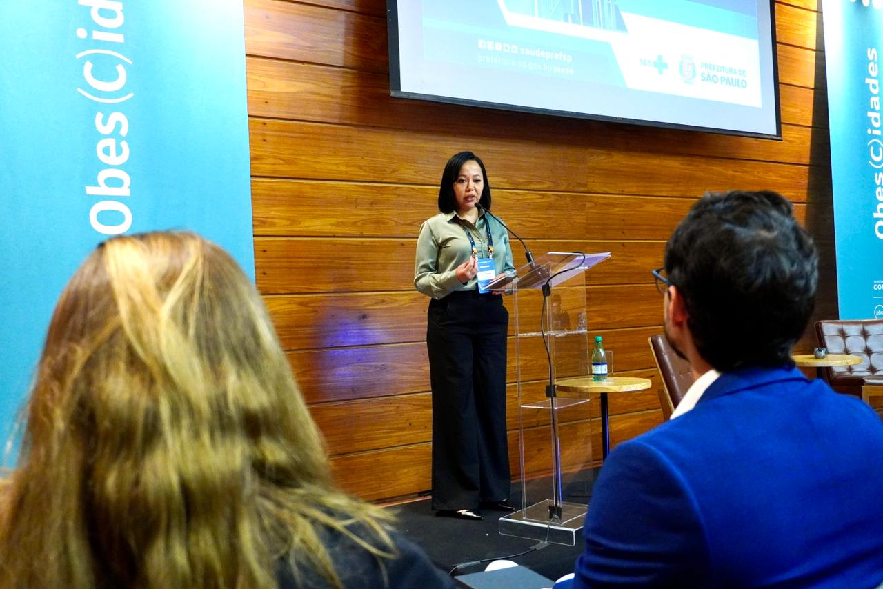 Uma mulher fala em pé atrás de um púlpito transparente, em um auditório de madeira clara. Ela é a nustricionista Josie Miranda que veste camisa verde-clara e calça preta, e segura o microfone enquanto se dirige ao público. À frente dela, duas pessoas estão sentadas, uma de cabelos loiros e outra de terno azul. Atrás da palestrante, há uma tela de projeção com o logotipo da Prefeitura de São Paulo e do SUS, e faixas verticais azuis com o texto “Obes(C)idades”. Sobre o púlpito, há uma garrafa de água e um crachá pendurado no pescoço da oradora.