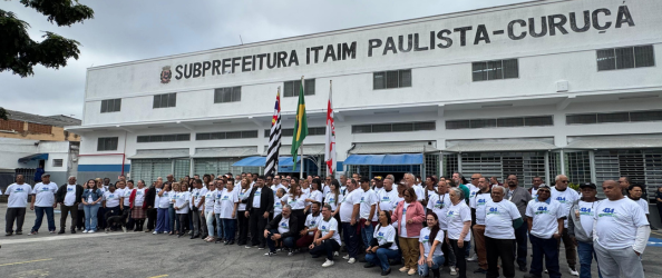 A imagem mostra um grande grupo de pessoas reunidas em frente ao prédio da Subprefeitura Itaim Paulista – Curuçá, em São Paulo. A fachada do prédio é branca, com janelas retangulares e a identificação da subprefeitura escrita em letras grandes e pretas no alto. No centro da imagem, à frente do grupo, estão hasteadas três bandeiras: a do Brasil, do estado de São Paulo e a da cidade de São Paulo. A maioria das pessoas está vestindo camisetas brancas com o logo de comemoração dos 414 anos do Itaim Paulista. O clima parece nublado, mas a iluminação é clara. A imagem transmite um momento de união e engajamento da comunidade em frente a um órgão público municipal.