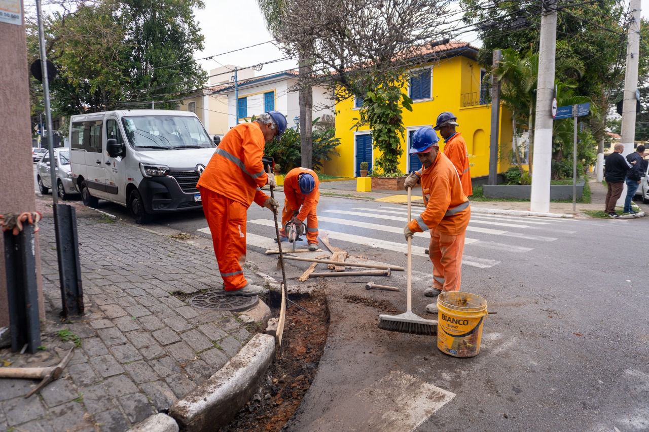 Foto 1: Trabalhador com uniforme laranja, capacete branco e luvas verdes realiza manutenção em bueiro, utilizando uma barra de ferro para o serviço. Foto 2: Linha de policiais militares perfilados ao lado de viaturas, com uma igreja amarela ao fundo em cenário urbano. Foto 3: Grupo de profissionais da saúde com jalecos brancos sorriem em frente a uma tenda da Subprefeitura de Santo Amaro, com mesa contendo vacinas, caixa de descarte e materiais médicos. Foto 4: Trabalhadores da prefeitura, com uniformes laranja e capacetes azuis, realizam manutenção no asfalto em uma esquina de bairro residencial. Foto 5: Grupo de policiais militares fardados, em formação, conversa com um homem de camisa azul em uma rua movimentada.