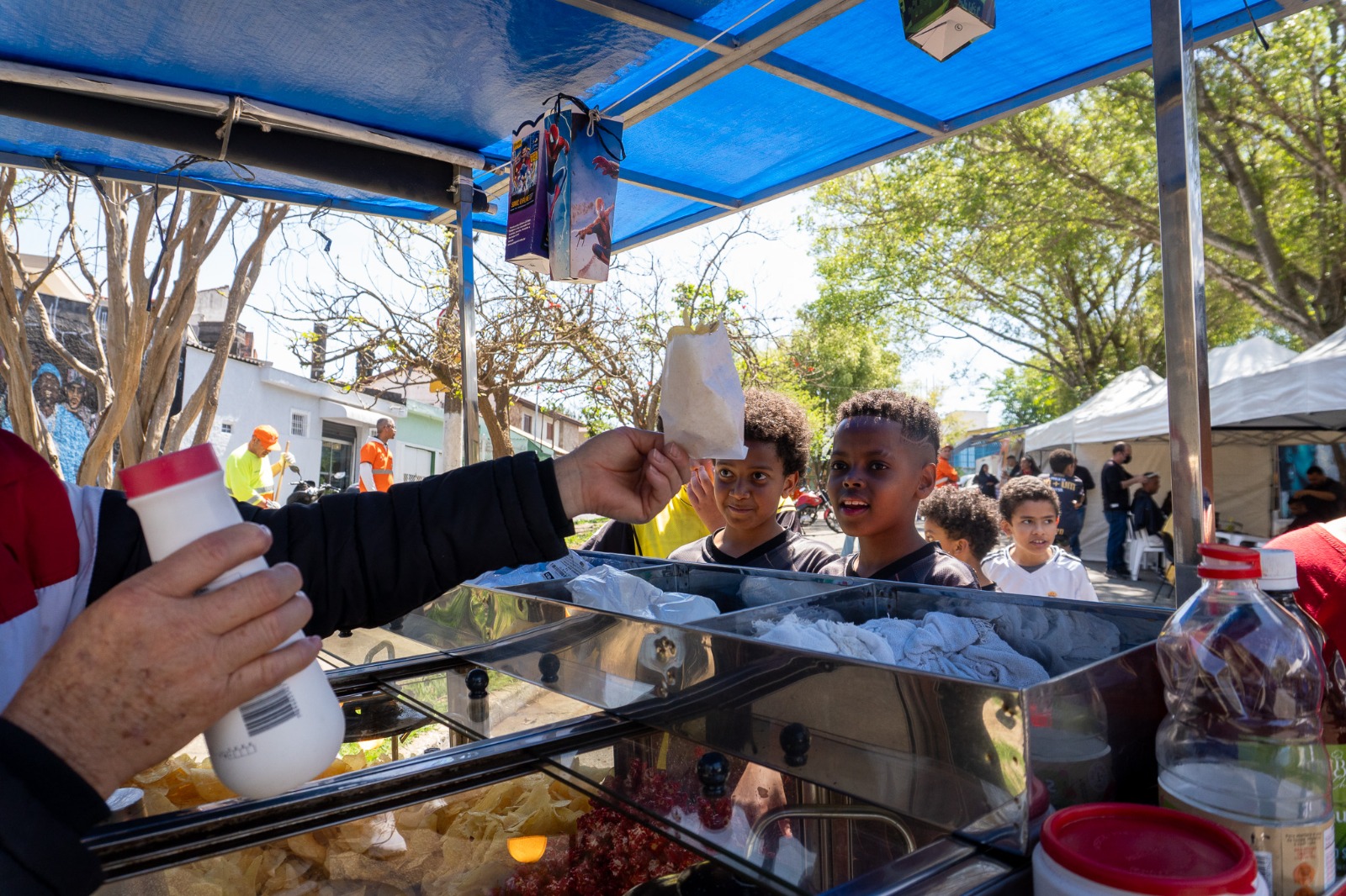 Primeira foto: Homem corta o cabelo de outro em ação comunitária ao ar livre, com o logo da subprefeitura de Santo Amaro ao fundo. Segunda foto: Crianças aguardam animadas para receber saquinhos de pipoca em barraca durante evento de rua.