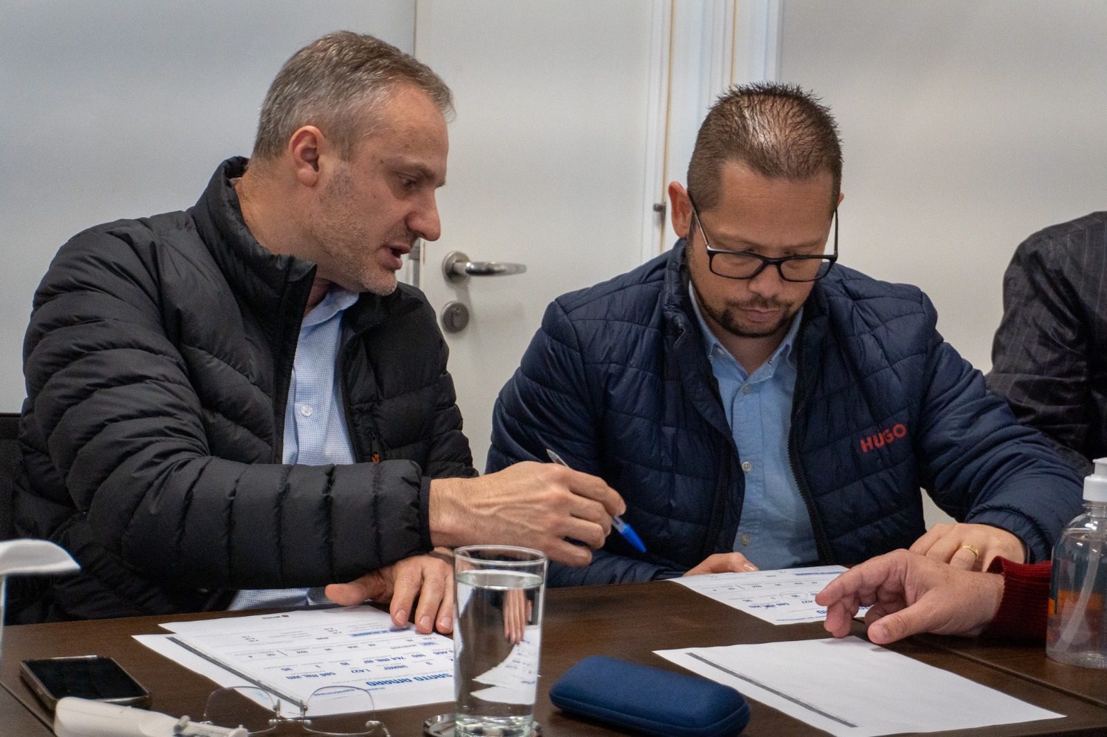 Dois homens sentados à mesa analisam documentos impressos. Um deles aponta para o papel enquanto o outro faz anotações com uma caneta. Ambos vestem jaquetas e parecem concentrados em uma reunião de trabalho.