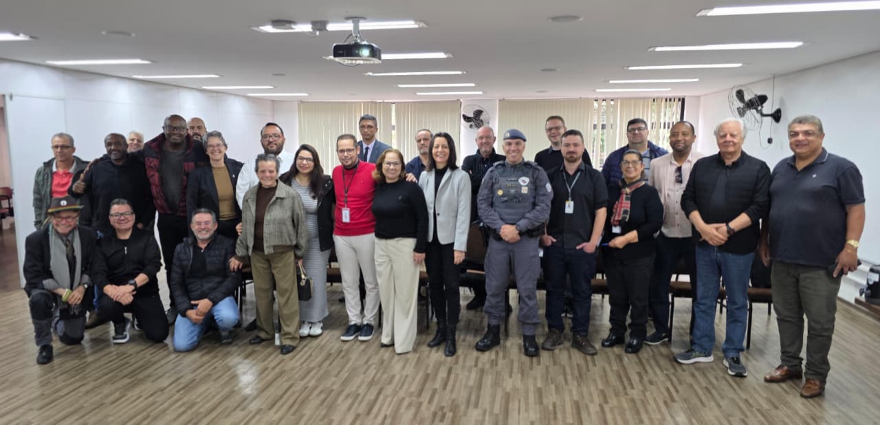 Descritivo das imagens: Primeira imagem: grupo de pessoas posa sorrindo em uma sala de reuniões, entre elas servidores públicos, representantes da Polícia Militar e lideranças locais. Segunda imagem: um policial fardado fala ao microfone durante reunião, enquanto outro homem com blusa vermelha e crachá o observa ao lado.