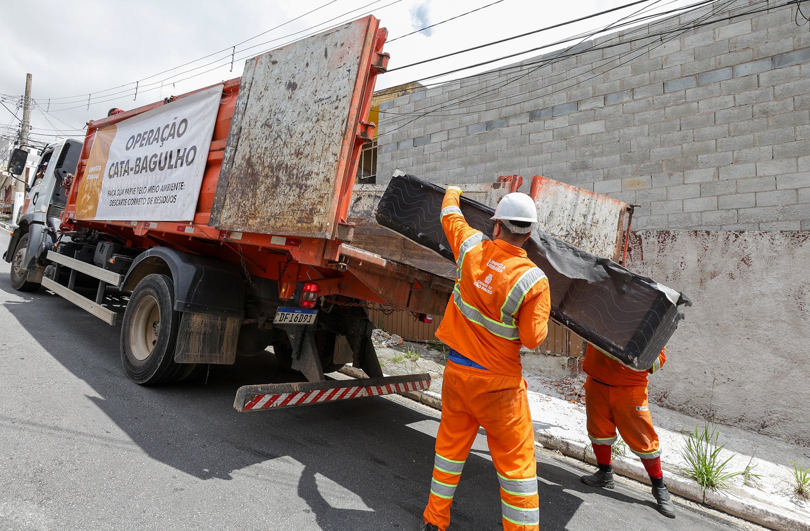 Na imagem, dois trabalhadores vestidos com uniformes laranja e faixas refletivas estão realizando a coleta de grandes objetos em uma rua. Eles estão colocando um colchão velho na caçamba de um caminhão de coleta. O caminhão é grande, de cor laranja e preta, e tem um cartaz lateral escrito “Operação Cata-Bagulho – Faça sua parte pelo meio ambiente. Descarte correto de resíduos”. Ambos os trabalhadores usam capacete branco e parecem estar em uma área urban