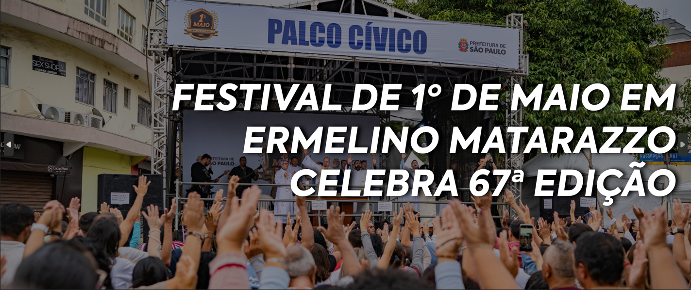 A imagem mostra uma fotografia panorâmica de um evento público ao ar livre durante o dia. Em primeiro plano, há uma grande multidão de pessoas vistas de costas, com os braços e as mãos levantados em direção ao palco.  No centro, há um palco montado com estrutura de metal (grid). Na parte superior do palco, uma faixa branca traz o texto em letras azuis: 