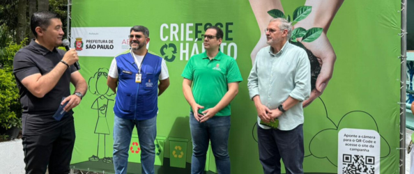 Em área externa, quatro homens estão diante de um painel verde da campanha “Crie Esse Hábito”. À esquerda, um homem fala ao microfone enquanto os outros observam. O painel mostra símbolos de reciclagem, uma muda de planta e um QR Code, indicando ação de conscientização ambiental.