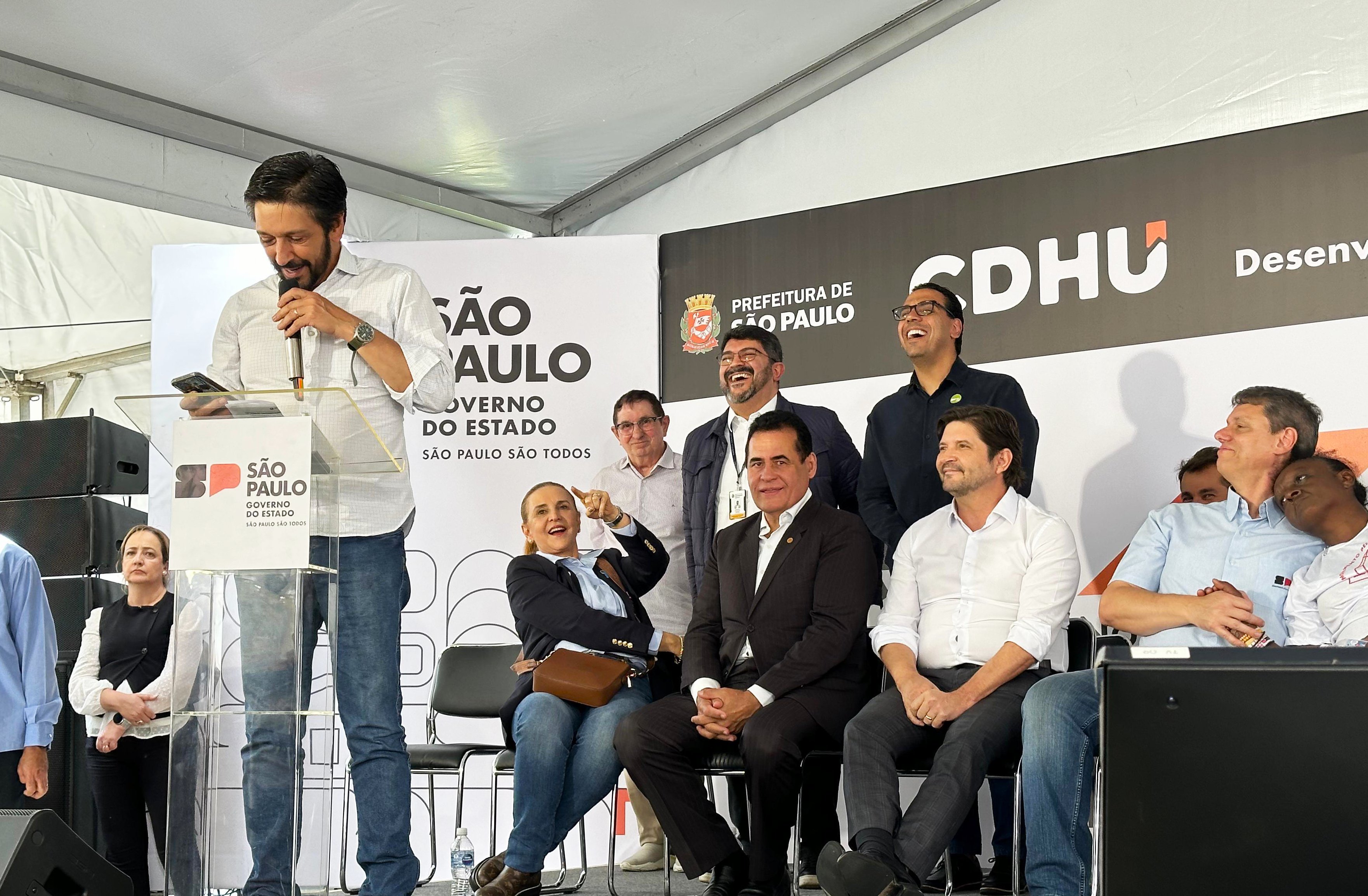 Em um palco coberto por uma tenda branca, várias pessoas participam de um evento oficial. À esquerda, o Prefeito de São Paulo, Ricardo Nunes, de camisa clara fala ao microfone em um púlpito transparente com o logotipo do Governo do Estado de São Paulo. Atrás e ao lado dele, outras pessoas estão sentadas e em pé, sorrindo e observando o discurso. No fundo, há painéis com os logotipos da Prefeitura de São Paulo e da CDHU (Companhia de Desenvolvimento Habitacional e Urbano). O clima é descontraído, com algumas pessoas rindo e interagindo entre si.