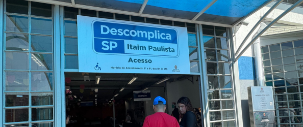 A imagem mostra a entrada da unidade do Descomplica SP – Itaim Paulista, um serviço da Prefeitura de São Paulo que oferece atendimentos públicos em um único local.  Acima da entrada, há uma placa com fundo branco e escrita em azul com os seguintes dizeres: 