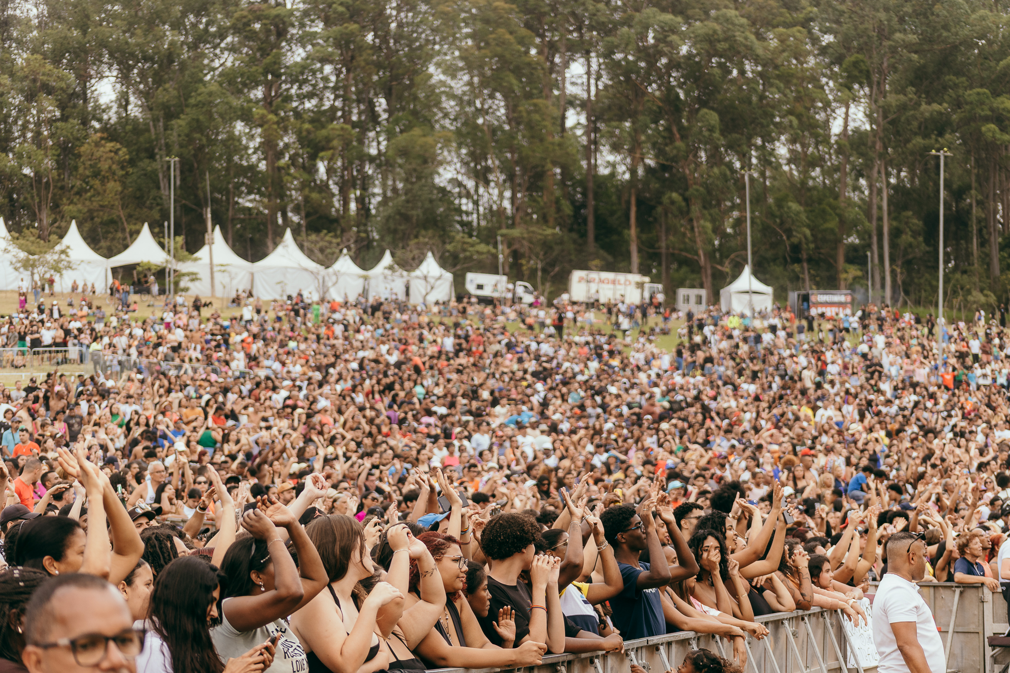 Multidão reunida ao ar livre durante um grande evento no Parque do Carmo. Na área frontal, muitas pessoas de diferentes idades, tons de pele e estilos levantam os braços, aplaudem e sorriem, demonstrando entusiasmo. Ao fundo, o gramado em declive está completamente tomado pelo público, formando uma grande massa de pessoas. Entre as árvores altas que cercam o espaço, há várias tendas brancas montadas para a programação. A cena transmite clima festivo, vibrante e de celebração coletiva.