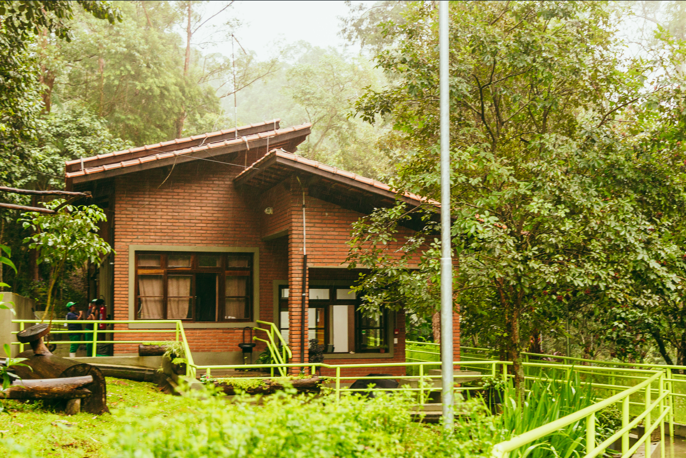 A imagem mostra uma construção térrea de tijolos aparentes localizada em uma área de mata bastante densa. O telhado da casa é de telhas cerâmicas inclinadas, com beirais salientes, e as esquadrias das janelas e portas são de madeira escura, com detalhes de pintura verde emoldurando as aberturas. Em volta da edificação, há árvores de médio porte e vegetação arbustiva que reforçam o clima de floresta. Na frente do prédio, uma rampa de acessibilidade com guarda-corpo pintado em verde vibrante acompanha a topografia do terreno, ligando o nível da rua até a entrada principal da construção. No canto esquerdo da foto, há uma escultura ou peça de madeira de grandes dimensões, com aparência rústica, posicionada sobre o gramado. Algumas pessoas com uniformes verdes podem ser vistas parcialmente atrás da vegetação, próximas à lateral da edificação. O tempo parece úmido e nublado, sugerindo que a foto foi tirada durante ou logo após uma chuva leve, pois é possível notar gotas de água e a presença de névoa entre as árvores no fundo da cena.