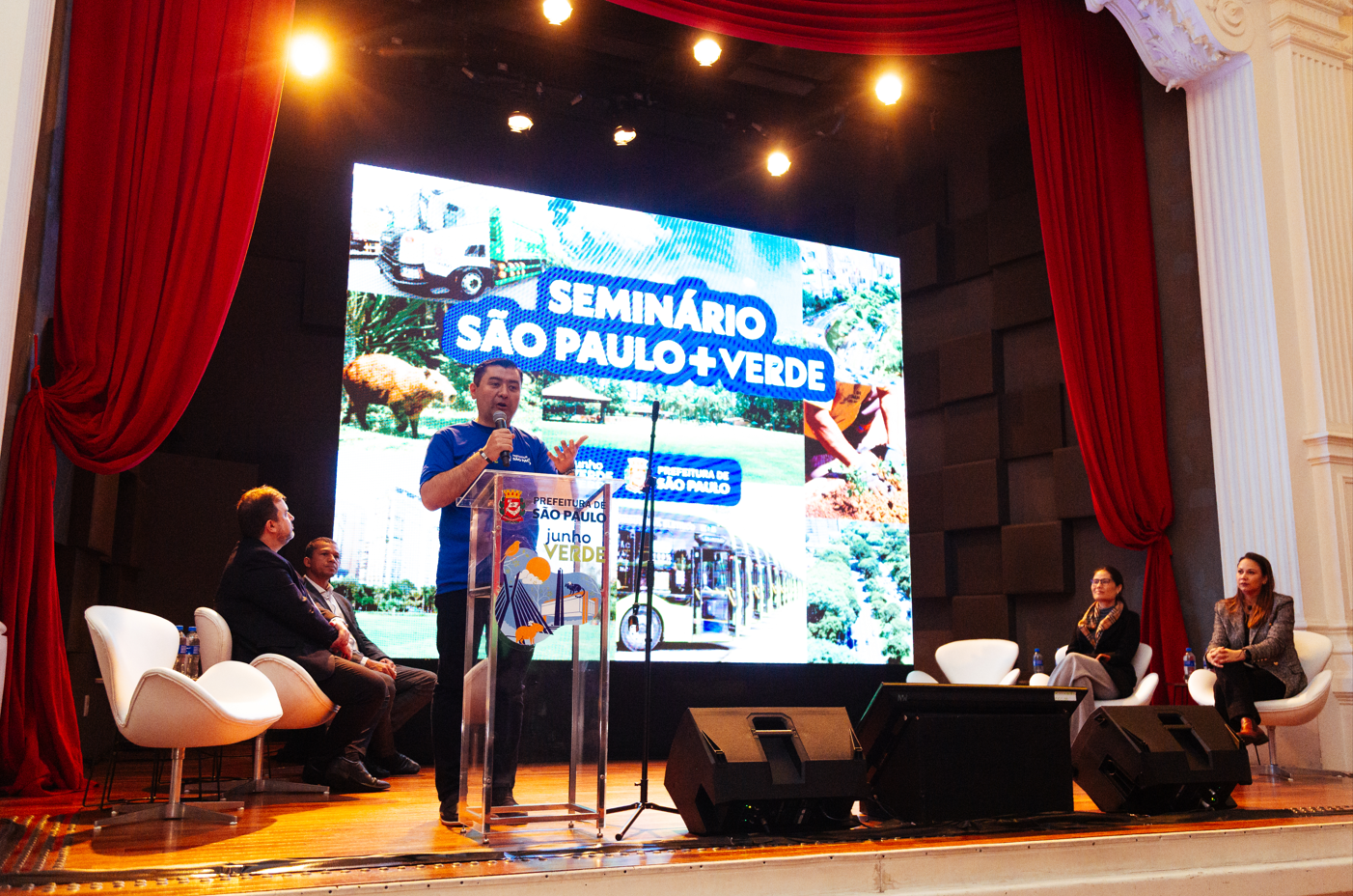 A imagem mostra uma cena do Seminário São Paulo + Verde, promovido pela Prefeitura de São Paulo. O evento ocorre em um auditório com cortinas vermelhas e arquitetura clássica. No centro do palco, um homem com camiseta azul da Prefeitura está discursando em um púlpito transparente, decorado com o selo “junho VERDE” — possivelmente em alusão ao mês dedicado à conscientização ambiental.  Ao fundo, um telão exibe imagens relacionadas ao meio ambiente urbano: um ônibus cercado de vegetação, uma capivara em área verde, plantio de mudas, além de um caminhão da limpeza urbana. À direita e à esquerda do orador, outras pessoas estão sentadas, sugerindo uma mesa de debate ou autoridades convidadas.  O ambiente e o material gráfico reforçam o compromisso com ações sustentáveis, como arborização, limpeza urbana, preservação da fauna e mobilidade verde — temas centrais do seminário.